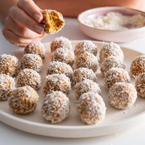 Hand holding a half-bitten Carrot Cake Bliss Ball with the plate in the background.