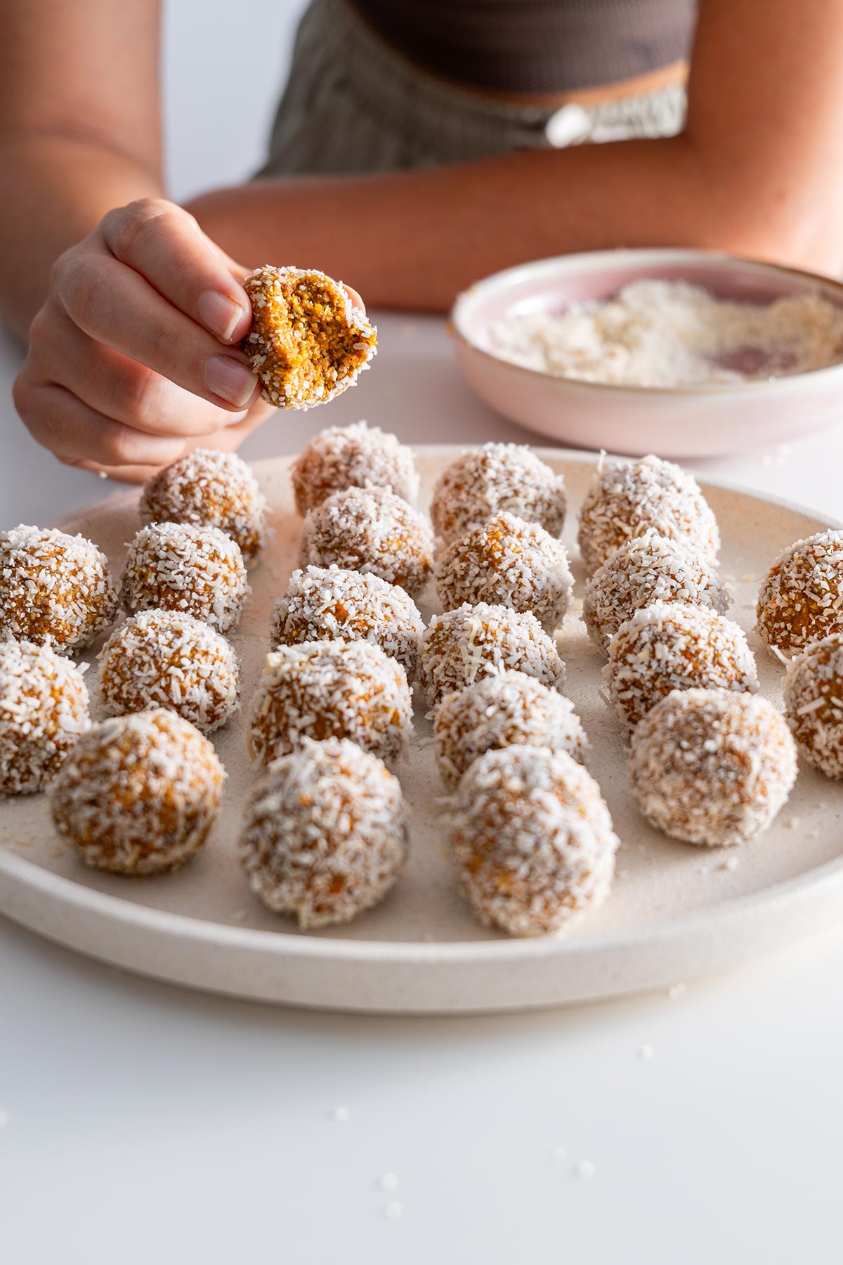 Hand holding a half-bitten Carrot Cake Bliss Ball with the plate in the background.