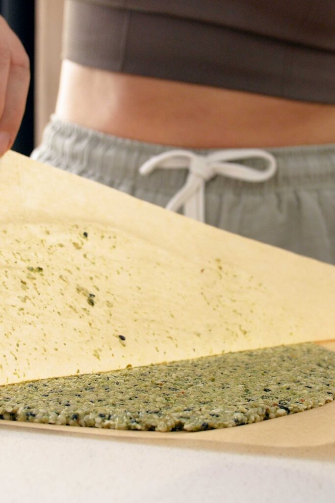 Top layer of baking paper being removed to reveal flatenned pumpkin seed cracker mix