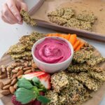 Wooden serving board with a vibrant pink beetroot hummus in the centre and healthy pumpkin seed crackers and other nibbles surrounding it