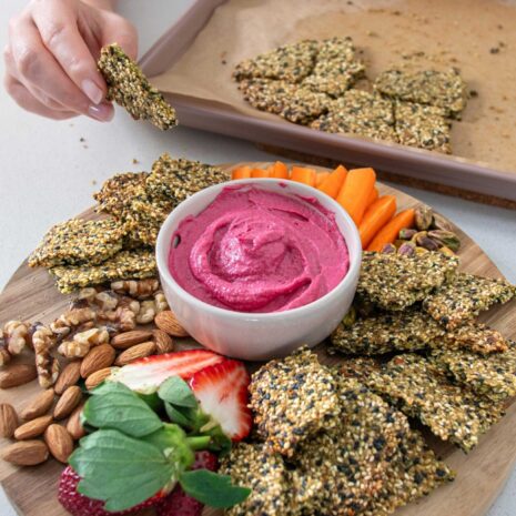 Wooden serving board with a vibrant pink beetroot hummus in the centre and healthy pumpkin seed crackers and other nibbles surrounding it