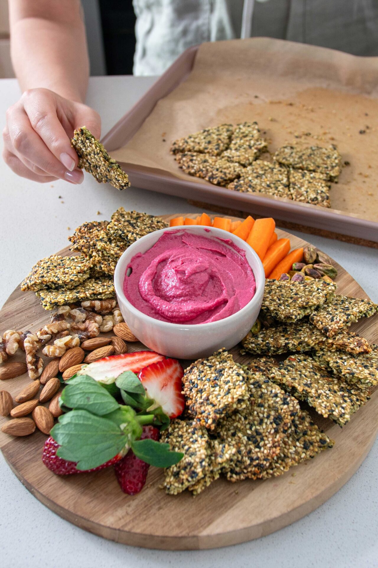 Wooden serving board with a vibrant pink beetroot hummus in the centre and healthy pumpkin seed crackers and other nibbles surrounding it