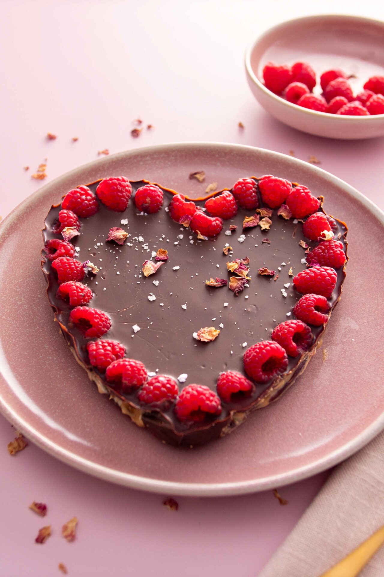 A heart shaped chocolate date bark with a border of vivid pink raspeberries on a pink background.