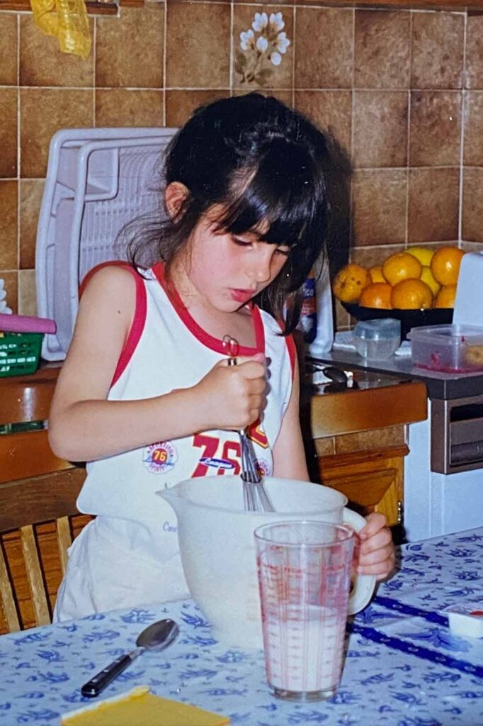 Sarah Cobacho as a young child in an old looking photo whisking a jug in the kitchen