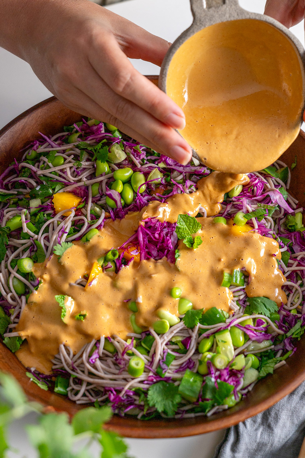Pouring peanut sauce over Soba Noodle Salad in a wooden bowl.