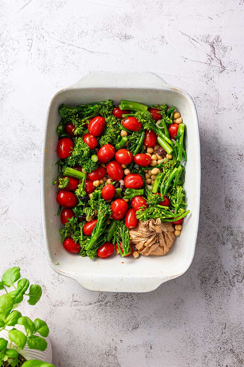 Roasted broccolini, cherry tomatoes, and chickpeas with an unpeeled garlic head in a white baking dish.