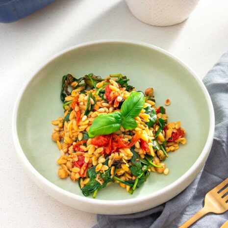 Plate of Mediterranean orzo salad with fresh spinach, tomatoes, and pine nuts, served with a gold fork and gray napkin.