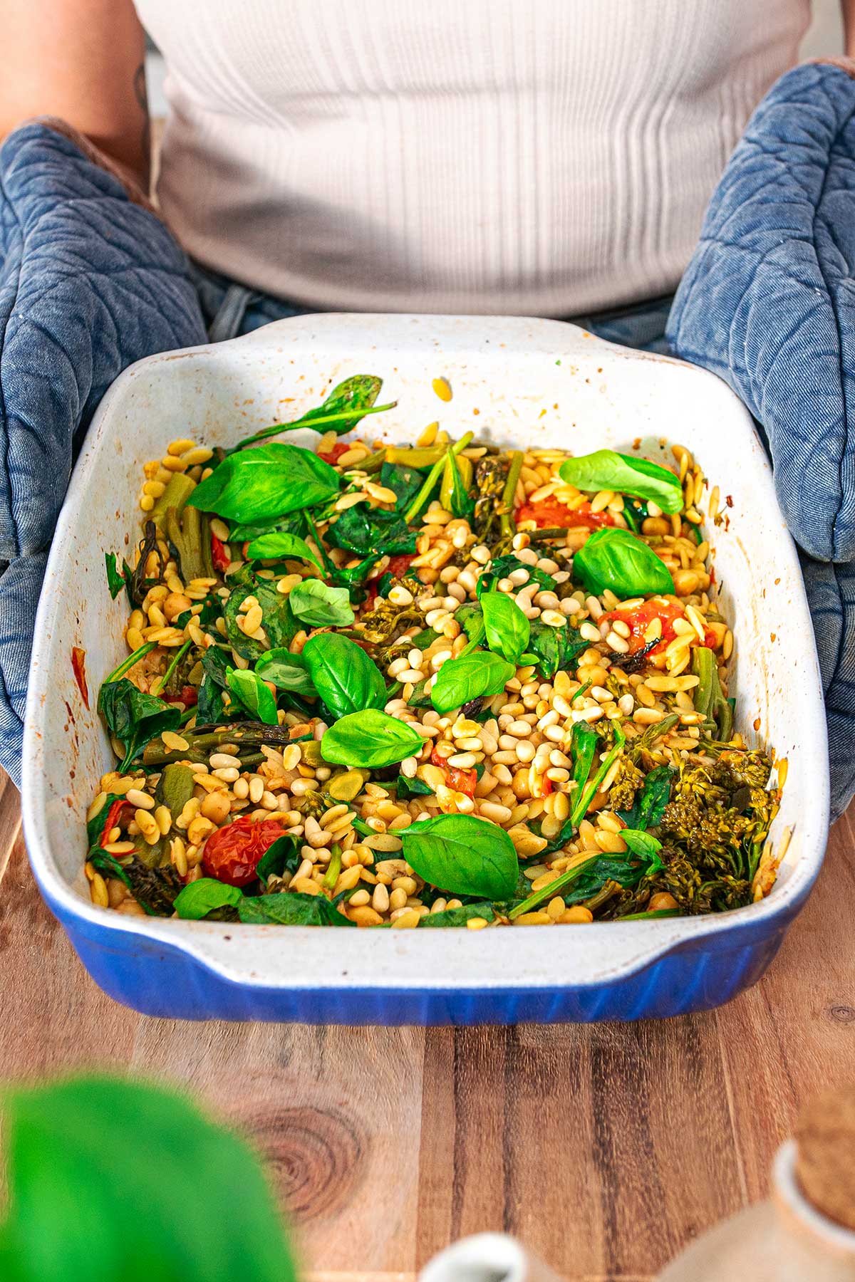 Orzo, spinach, cherry tomatoes, and pine nuts garnished with fresh basil in a ceramic baking dish being held by Sarah Cobacho with blue oven mittens
