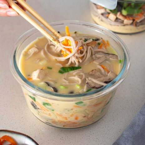 A close-up of high-protein miso noodle soup in a glass meal prep container, with a focus on the soba noodles, tofu, and vibrant vegetables.