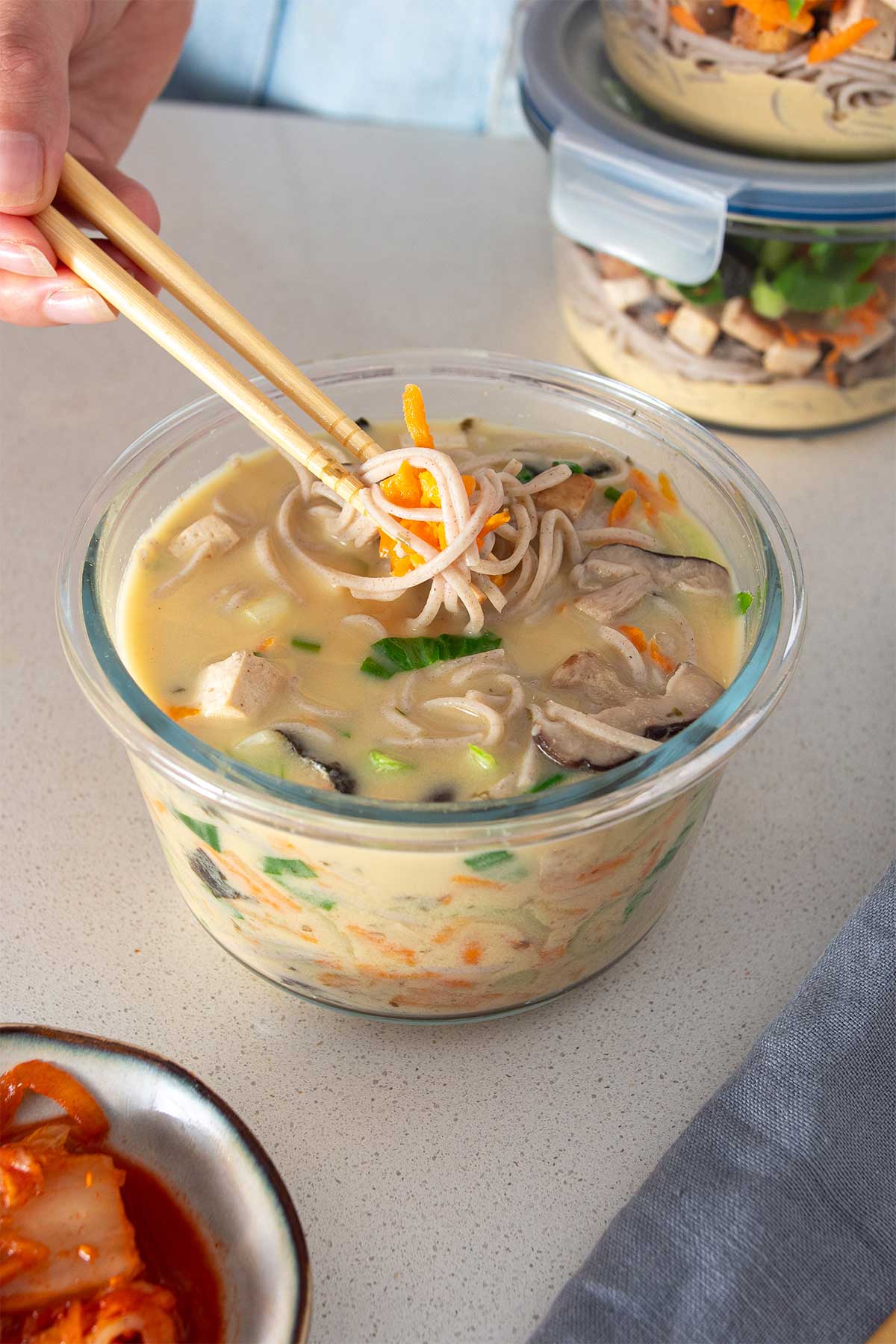 A close-up of high-protein miso noodle soup in a glass meal prep container, with a focus on the soba noodles, tofu, and vibrant vegetables.