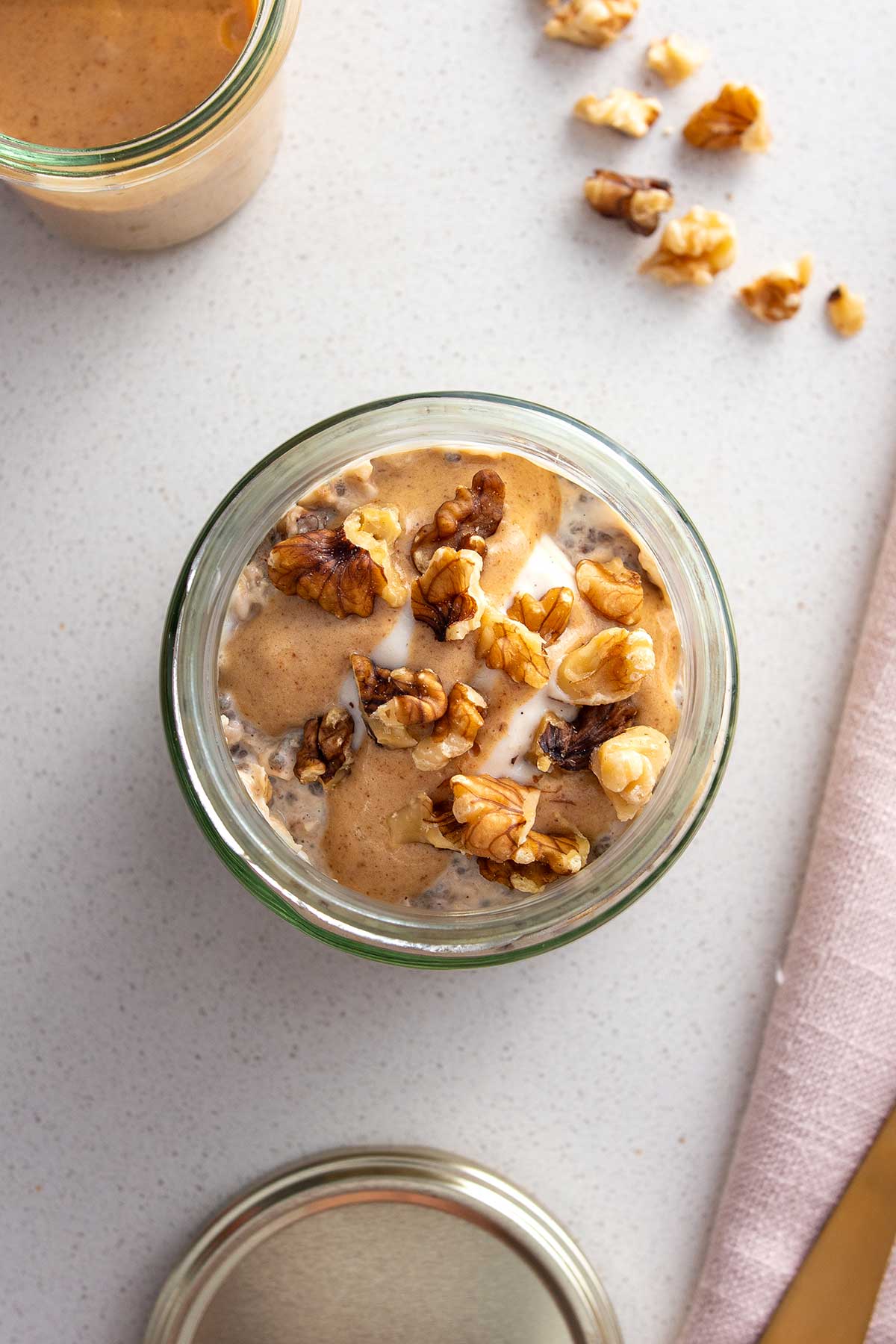 Top view of a jar of apple pie overnight oats topped with a drizzle of caramel sauce and a sprinkle of walnuts, set on a clean white countertop.