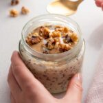 A hand holding a jar filled with apple pie overnight oats, garnished with crushed walnuts on top, ready to be enjoyed with a golden spoon resting beside it.