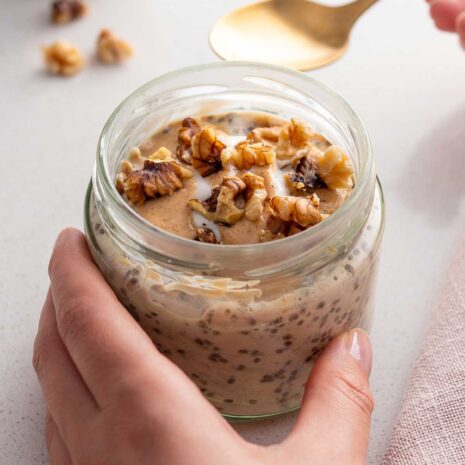 A hand holding a jar filled with apple pie overnight oats, garnished with crushed walnuts on top, ready to be enjoyed with a golden spoon resting beside it.