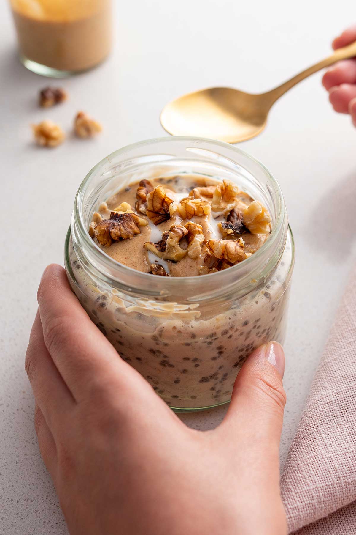 A hand holding a jar filled with apple pie overnight oats, garnished with crushed walnuts on top, ready to be enjoyed with a golden spoon resting beside it.