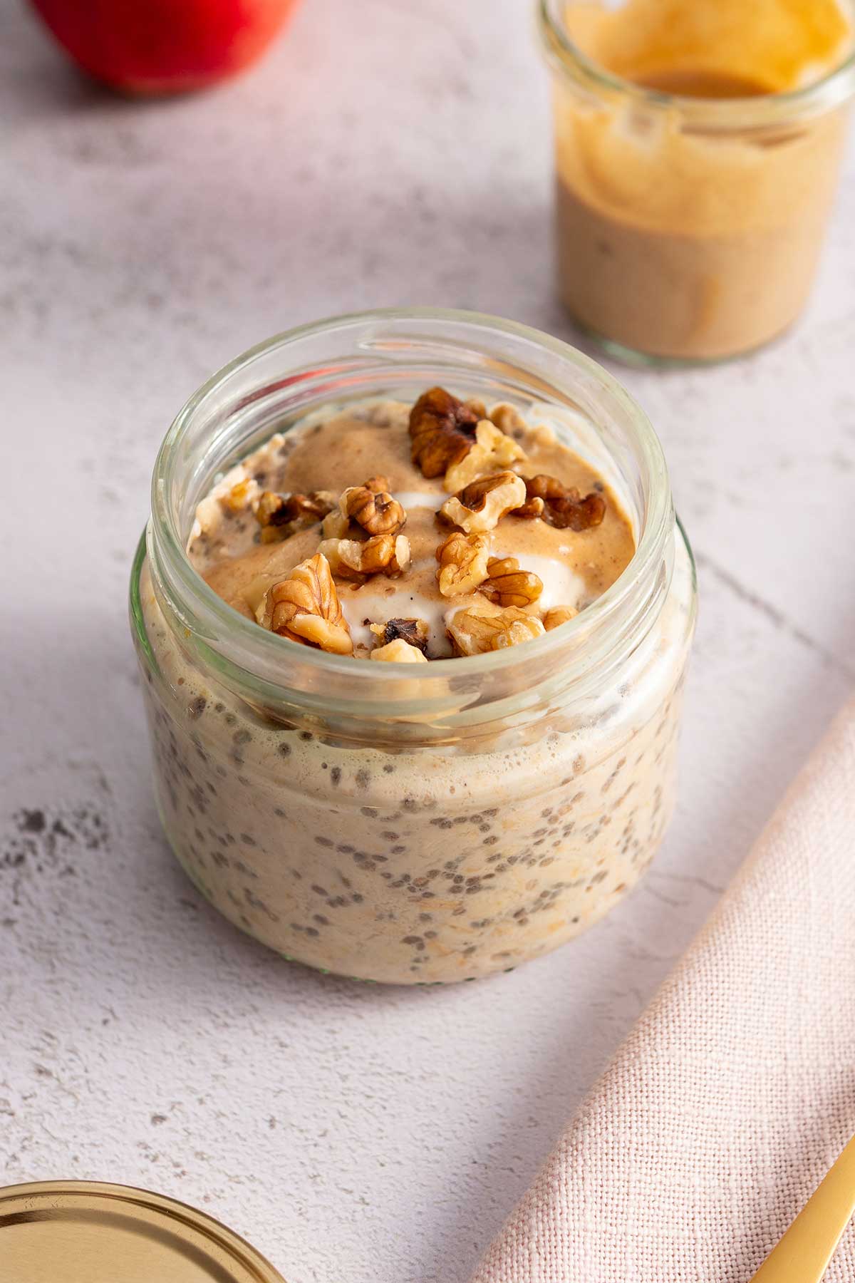 Close-up of a creamy jar of apple pie overnight oats topped with walnuts and caramel sauce, with a fresh apple and a jar of caramel in the background.