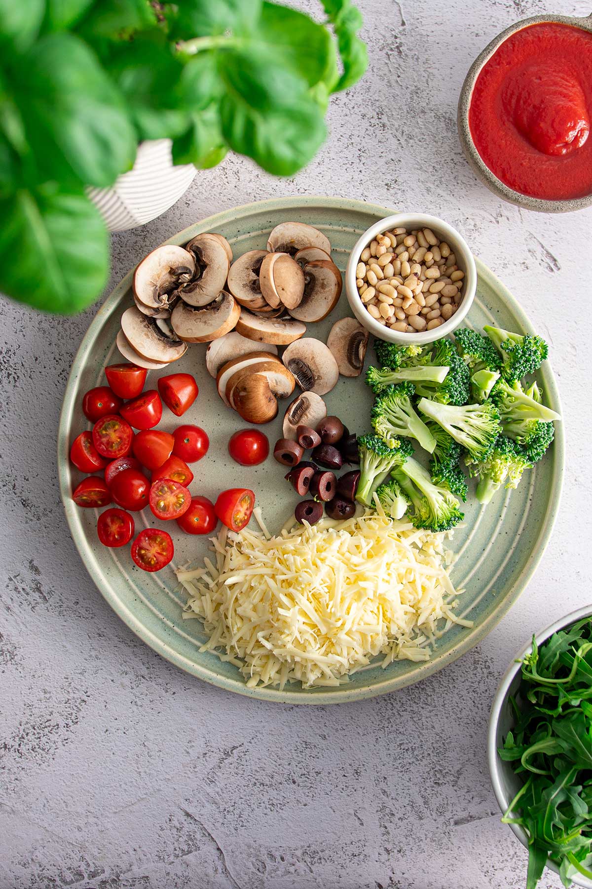A plate with neatly arranged pizza toppings including sliced mushrooms, cherry tomatoes, olives, broccoli, pine nuts, and shredded vegan cheese, all set for assembling on the pizza crust.
