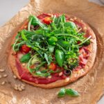 A completed gluten-free pizza with an array of colorful toppings including tomatoes, olives, greens, and broccoli, ready to be enjoyed, displayed on parchment paper against a modern kitchen backdrop.