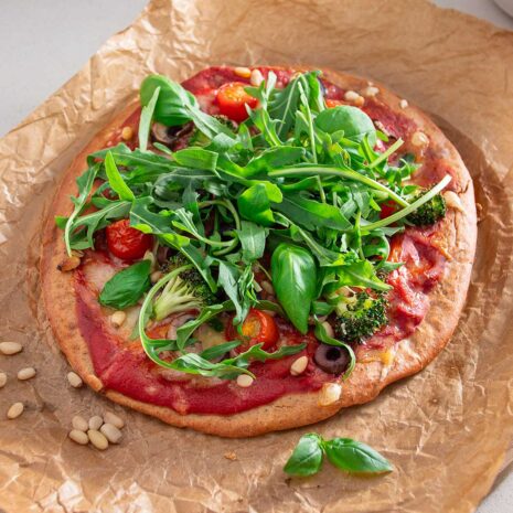 A completed gluten-free pizza with an array of colorful toppings including tomatoes, olives, greens, and broccoli, ready to be enjoyed, displayed on parchment paper against a modern kitchen backdrop.