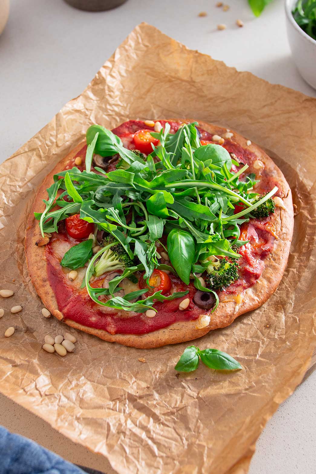 A completed gluten-free pizza with an array of colorful toppings including tomatoes, olives, greens, and broccoli, ready to be enjoyed, displayed on parchment paper against a modern kitchen backdrop.
