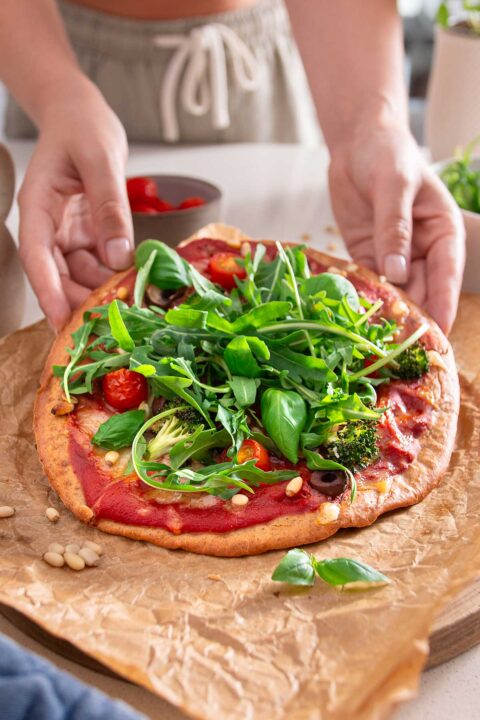 Hands delicately placing fresh arugula and herbs on a baked gluten-free pizza crust, showcasing the process of adding fresh, leafy greens to enhance the flavors of the vegan pizza.