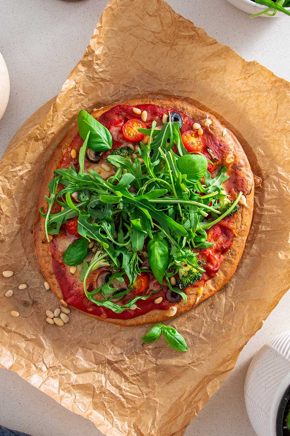 A freshly baked gluten-free pizza topped with a vibrant selection of greens, cherry tomatoes, olives, and pine nuts on a rustic brown baking paper, indicating a nutritious and delicious meal option.