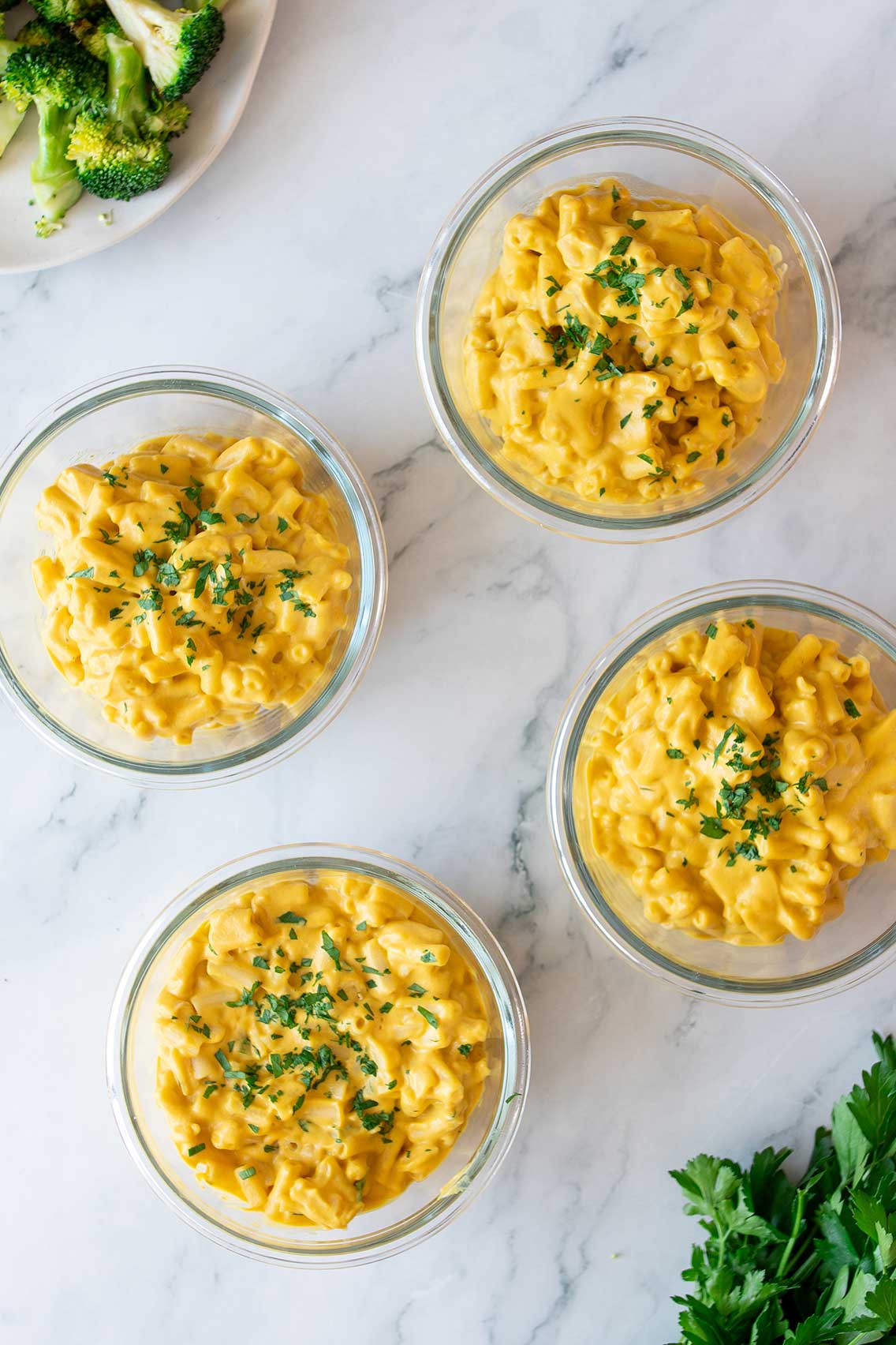 An overhead shot of four glass bowls containing vegan high-protein mac and cheese on a marble background, accompanied by fresh parsley.
