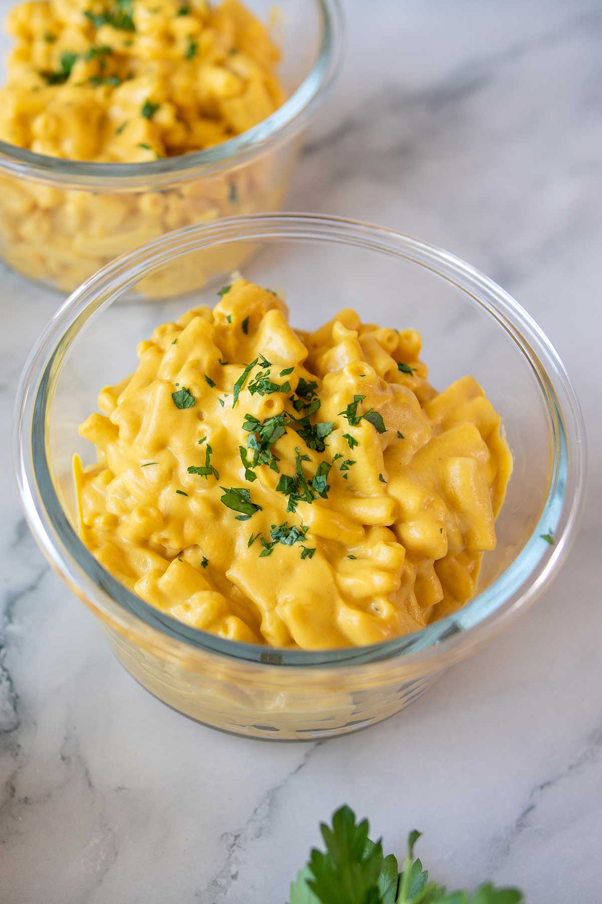 Two glass bowls on a marble counter filled with creamy vegan high-protein mac and cheese, topped with chopped fresh parsley.