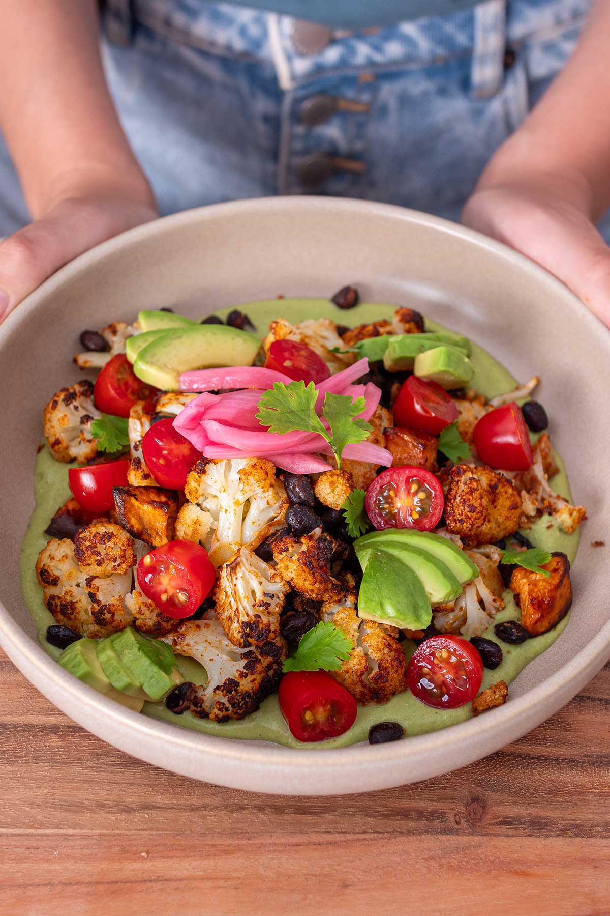 Sarah Cobacho presenting a bowl of roasted sweet potato and cauliflower in creamy cilantro sauce, garnished with avocado slices, pickled onions, and cherry tomatoes.