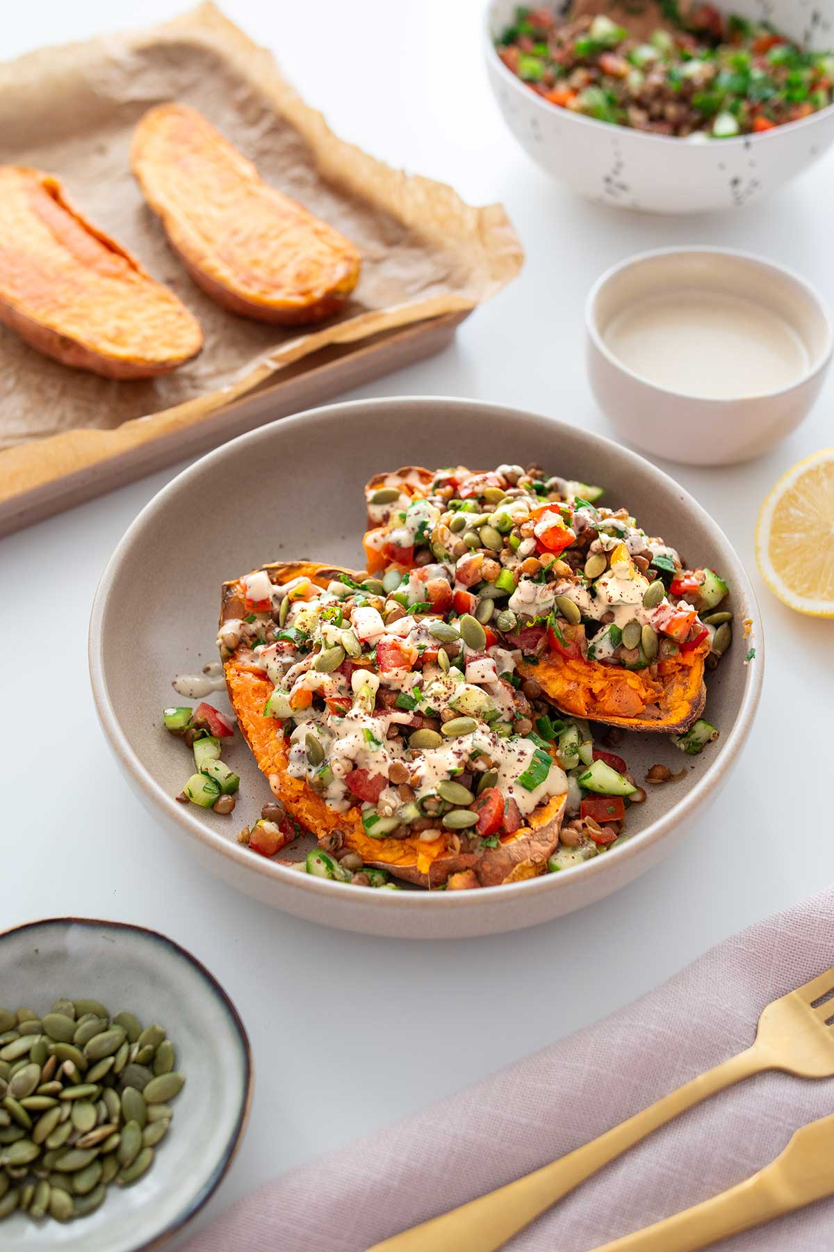 Roasted Sweet Potato Tabbouleh Bowl garnished with seeds and dressing, surrounded by roasted sweet potatoes on a baking tray and toppings.