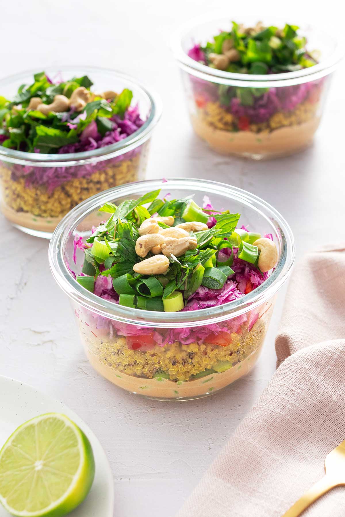 Close-up of a single glass meal prep container with layers of a nutritious quinoa salad, showing vibrant colors from the almond butter dressing, yellow turmeric quinoa, green edamame, red bell pepper, and purple cabbage topped with green herbs and cashews.