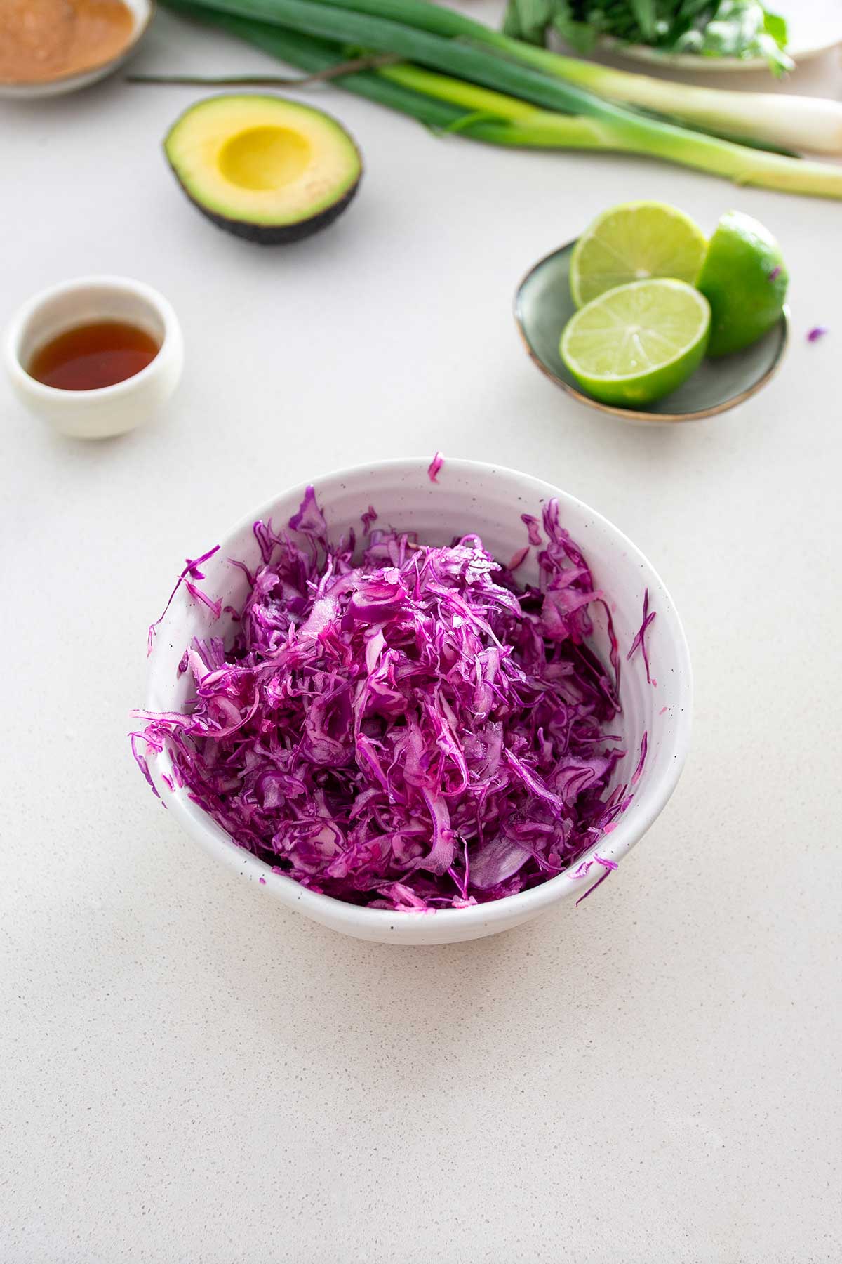 A white bowl on a kitchen countertop containing shredded red cabbage prepared for salad, with half an avocado, sliced limes, and a small bowl of maple syrup on the side, ready for recipe assembly.