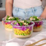 Three glass meal prep containers on a kitchen counter filled with layers of almond butter dressing, turmeric quinoa, edamame, shredded red cabbage, and topped with fresh green scallions, mint, and cashews.