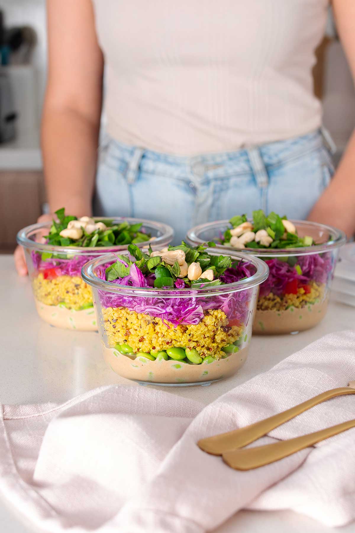 Three glass meal prep containers on a kitchen counter filled with layers of almond butter dressing, turmeric quinoa, edamame, shredded red cabbage, and topped with fresh green scallions, mint, and cashews.