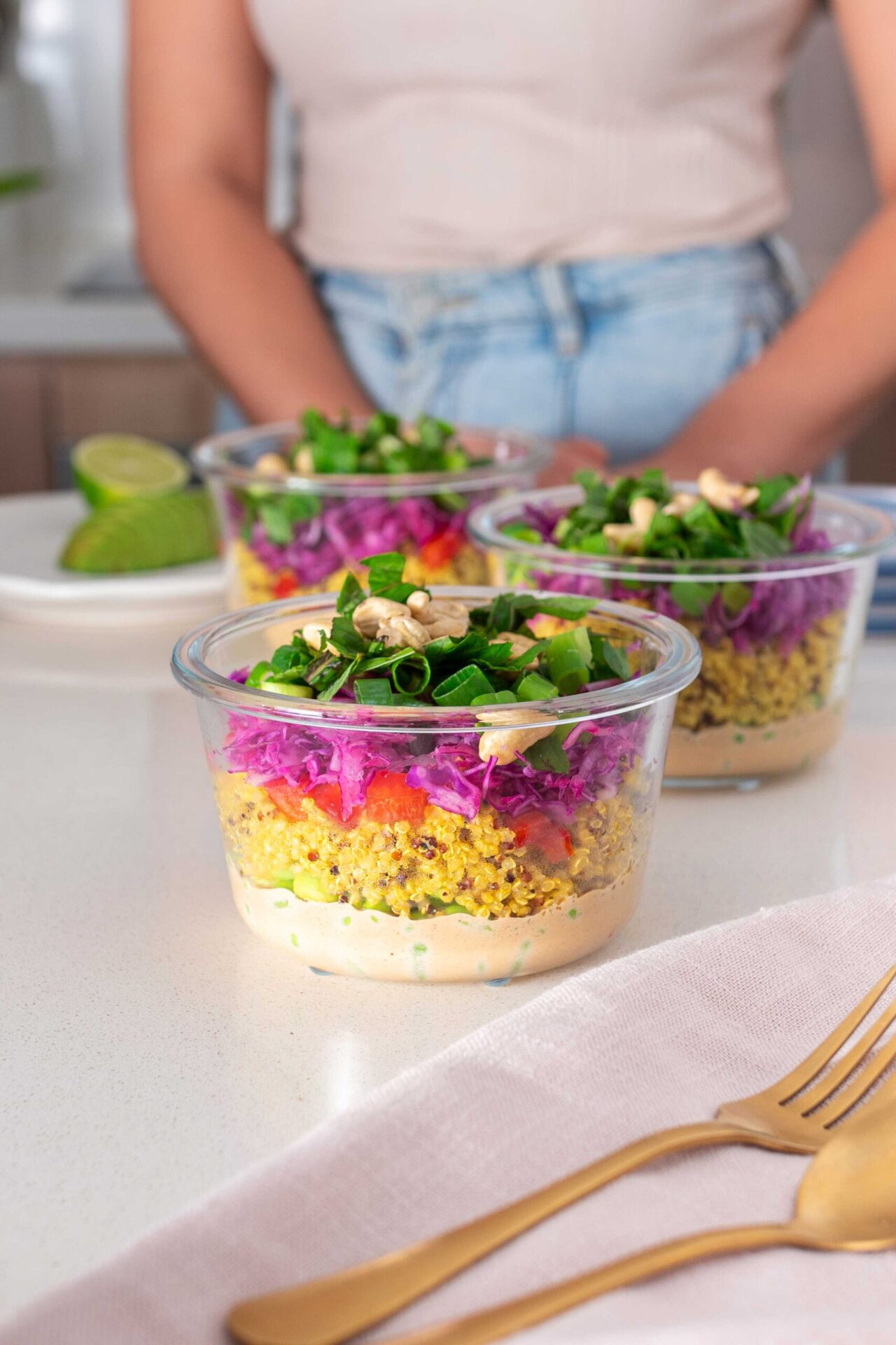 Three glass meal prep containers on a kitchen counter filled with layers of almond butter dressing, turmeric quinoa, edamame, shredded red cabbage, and topped with fresh green scallions, mint, and cashews, with a side of sliced lime and avocado.