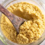 Close up photo of a glass jar filled with vegan parmesan being stirred by a wooden spoon