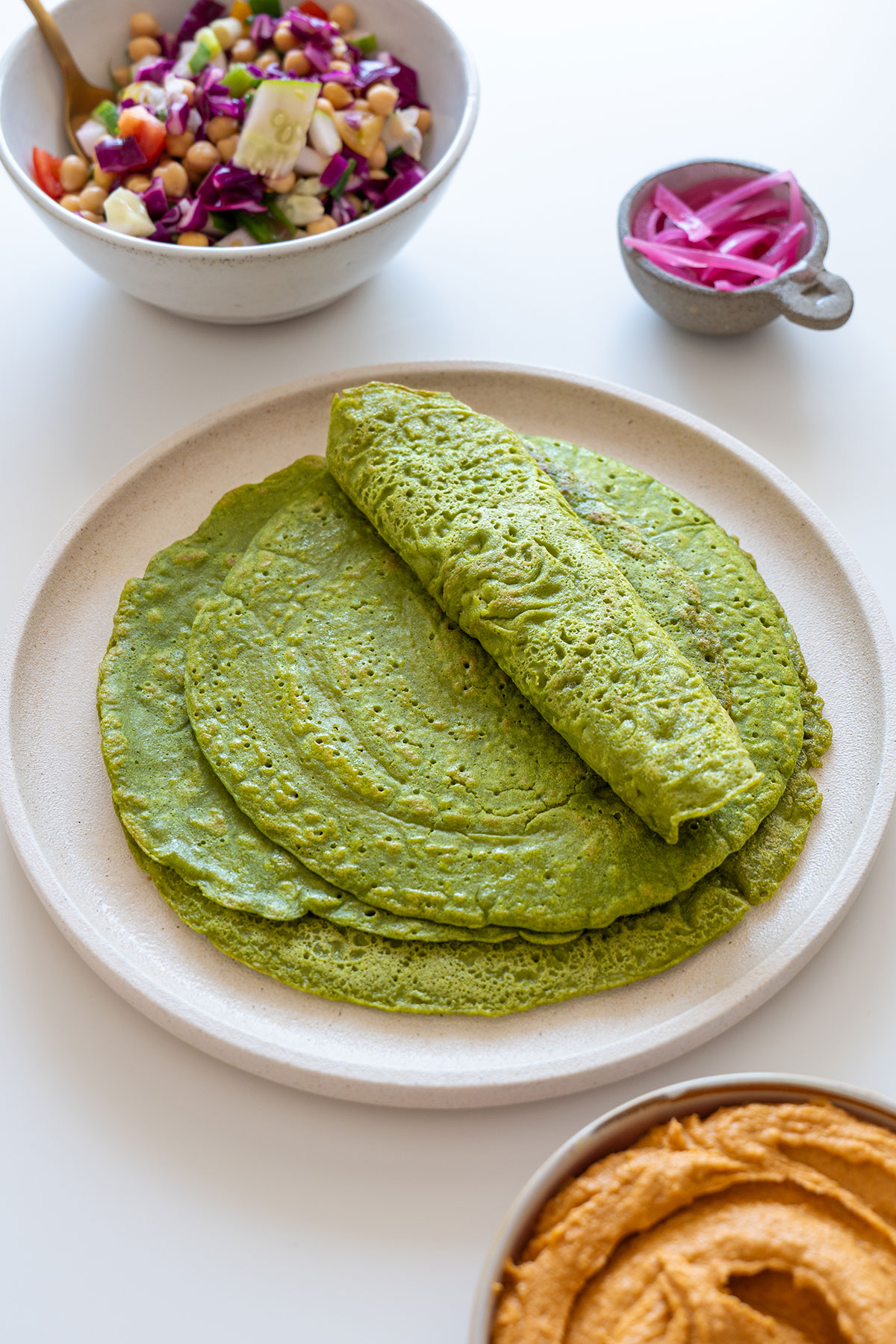 Overhead image of soft, green buckwheat spinach wraps stacked on a plate, with a colorful bowl of salad and dip in the background.