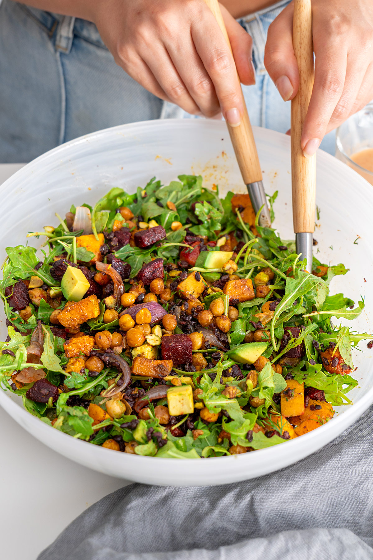 Hands tossing a Harissa Pumpkin Salad with arugula and avocado in a white bowl