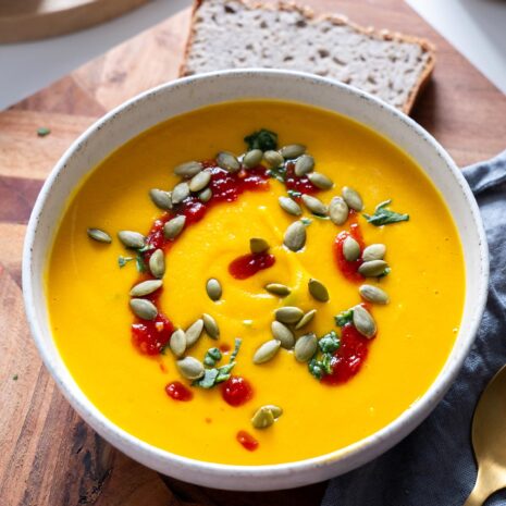 Closeup of a healing bowl of smooth butternut squash and red lentil soup garnished with seeds and red sauce, accompanied by slices of bread and a gold spoon on a rustic board.