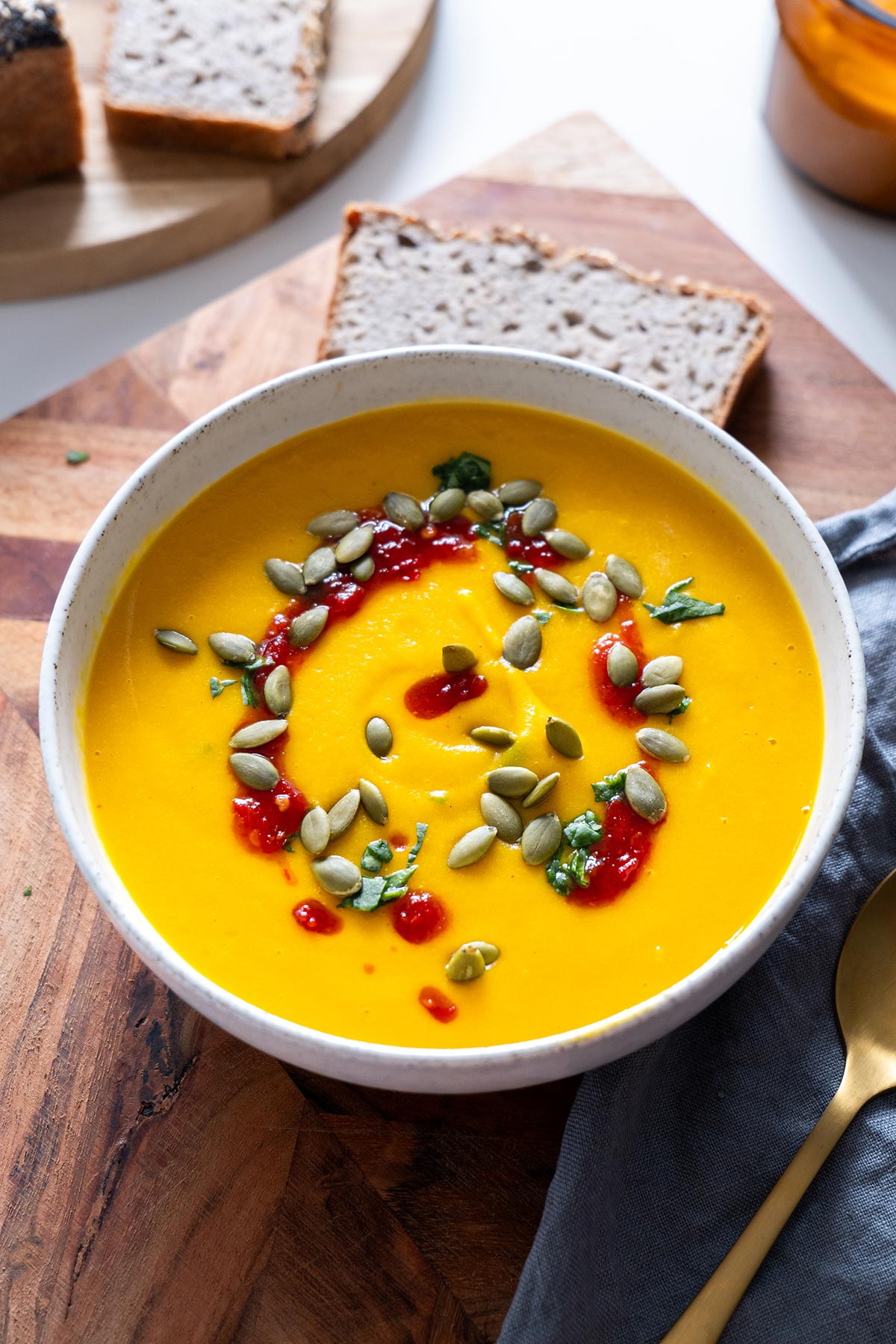 Closeup of a healing bowl of smooth butternut squash and red lentil soup garnished with seeds and red sauce, accompanied by slices of bread and a gold spoon on a rustic board.