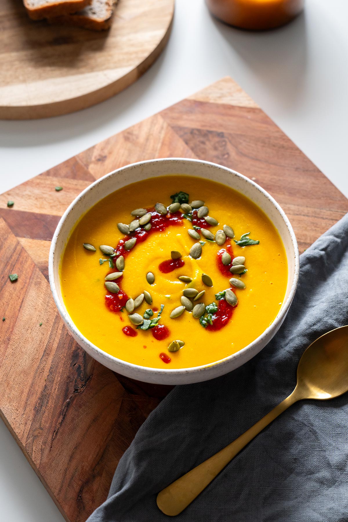 Creamy butternut squash and red lentil soup in a bowl on a wooden board with golden spoon. Topped with pumpkin seeds and a drizzle of red sauce, ready to be enjoyed.
