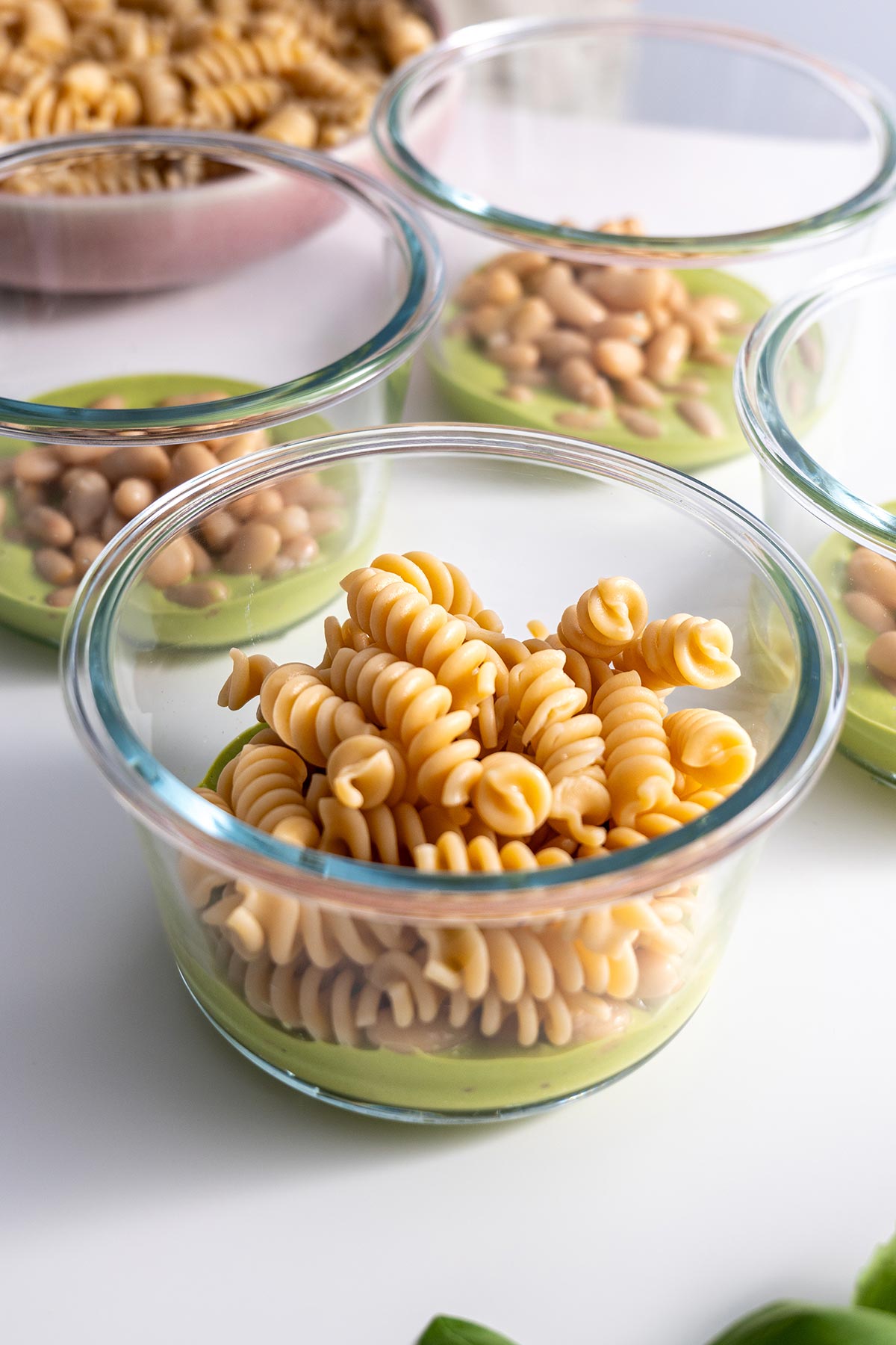 Step-by-step meal prep showing cooked spelt pasta portioned into four clear glass containers, ready for layering with other salad components.