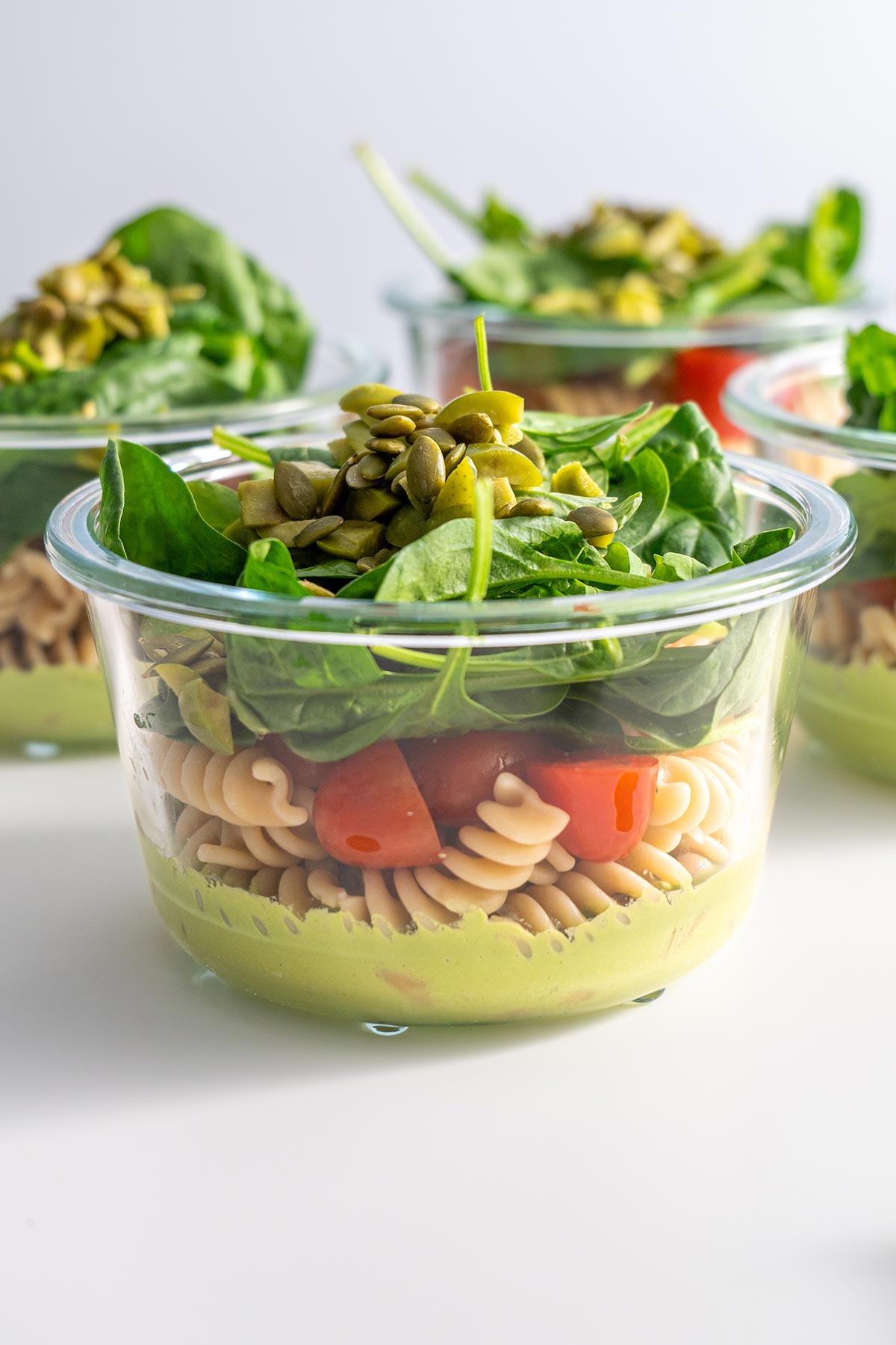 Progression of meal prep for Pistachio Pesto Pasta Salad, with the ingredients layered in glass containers; starting with a green pesto base, then adding beans, pasta, and tomatoes.