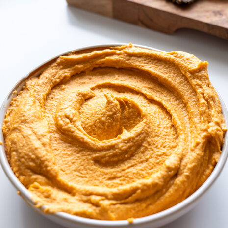 Close-up of a bowl of red lentil hummus with an enticing swirl pattern on top with crackers on a chopping board in the background.