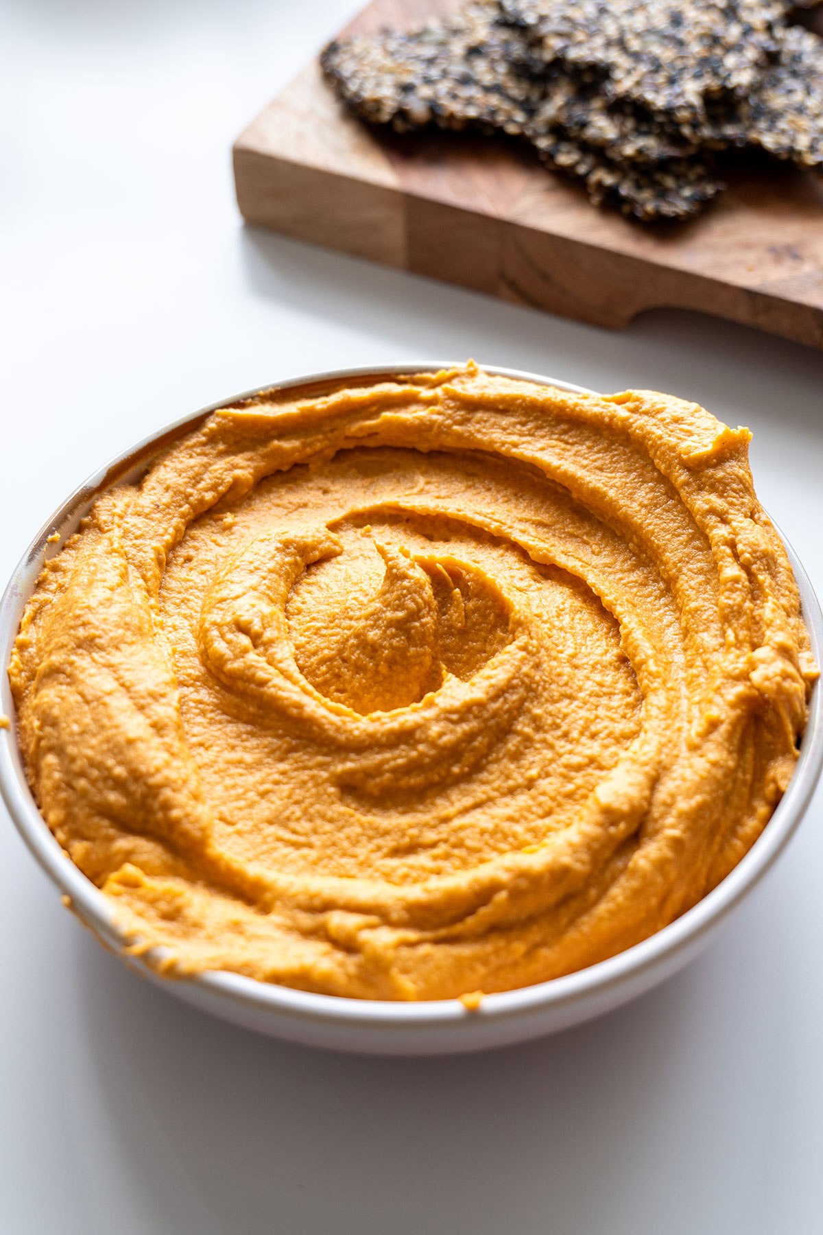 Close-up of a bowl of red lentil hummus with an enticing swirl pattern on top with crackers on a chopping board in the background.