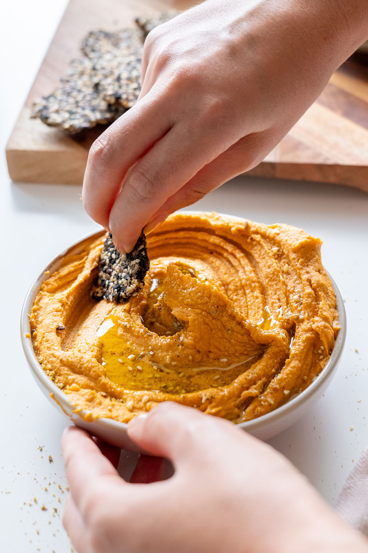 A hand dipping a black sesame cracker into a bowl of smooth red lentil hummus, ready to be enjoyed.