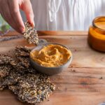 Hand holding a seeded cracker over a wooden table with hummus and a candle in the background.