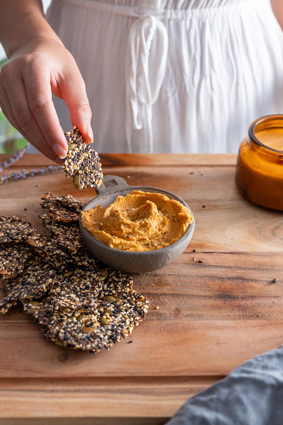 Hand holding a seeded cracker over a wooden table with hummus and a candle in the background.