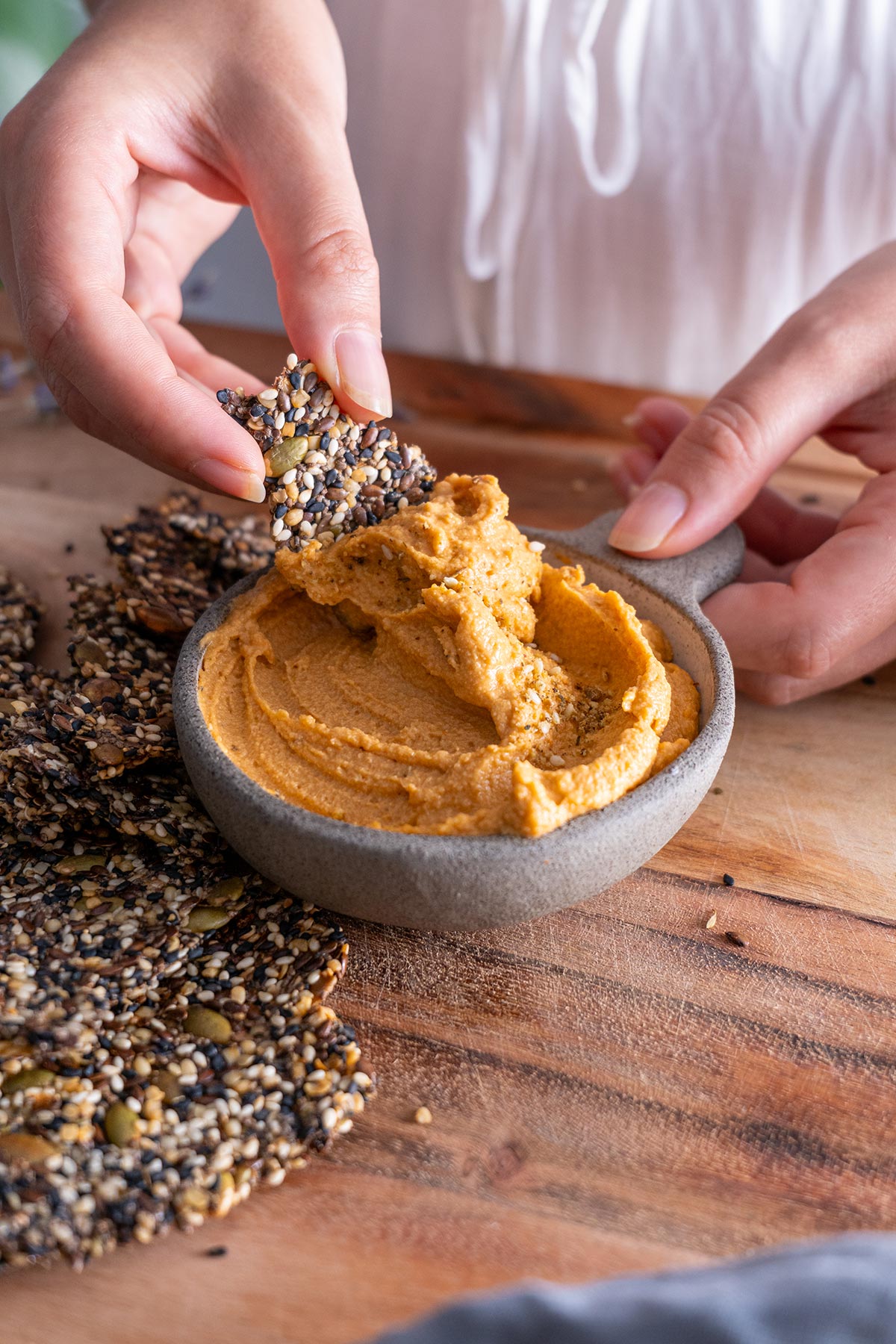 Closeup of a hand dipping a seeded cracker into red lentil hummus.