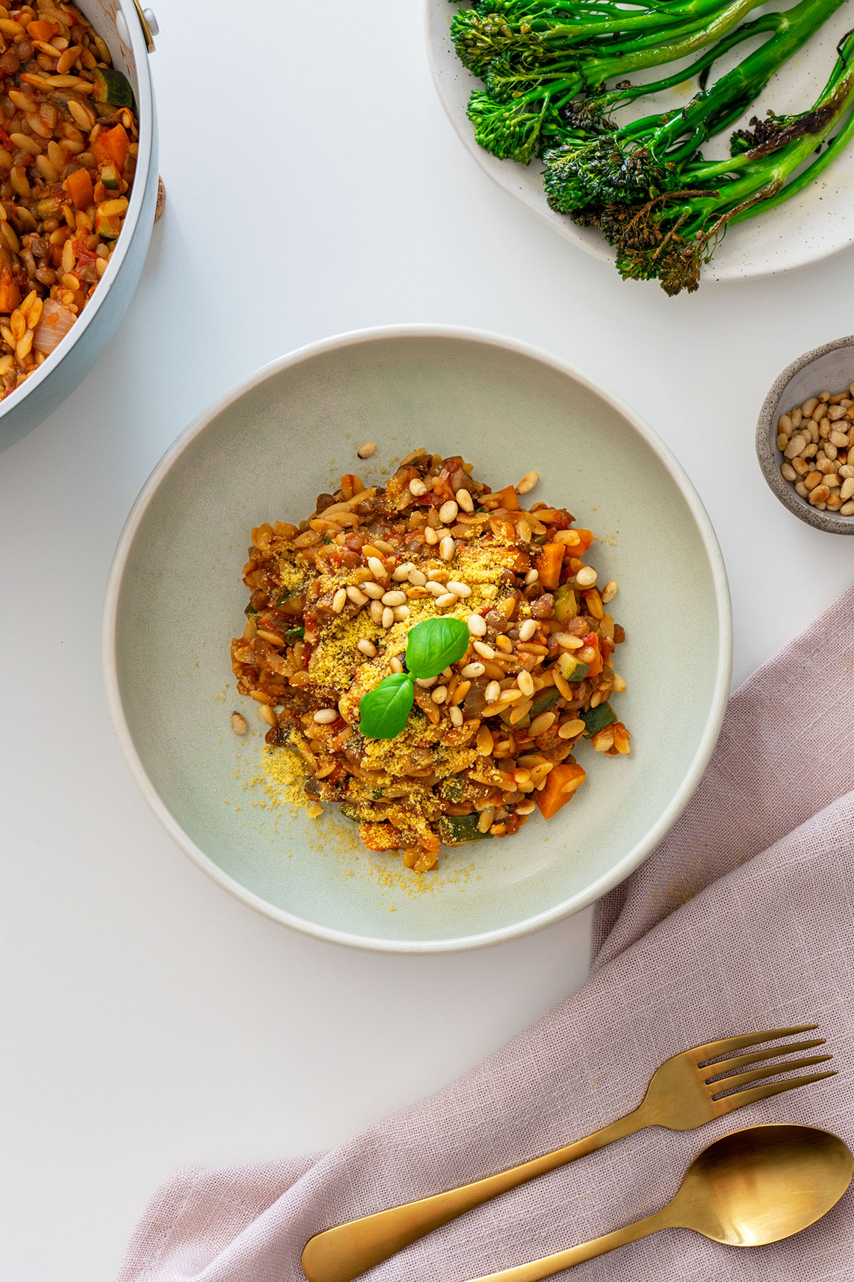 Overhead view of vegan one-pot orzo Bolognese in a bowl with a side of grilled broccolini and a pot of the meal in the background.
