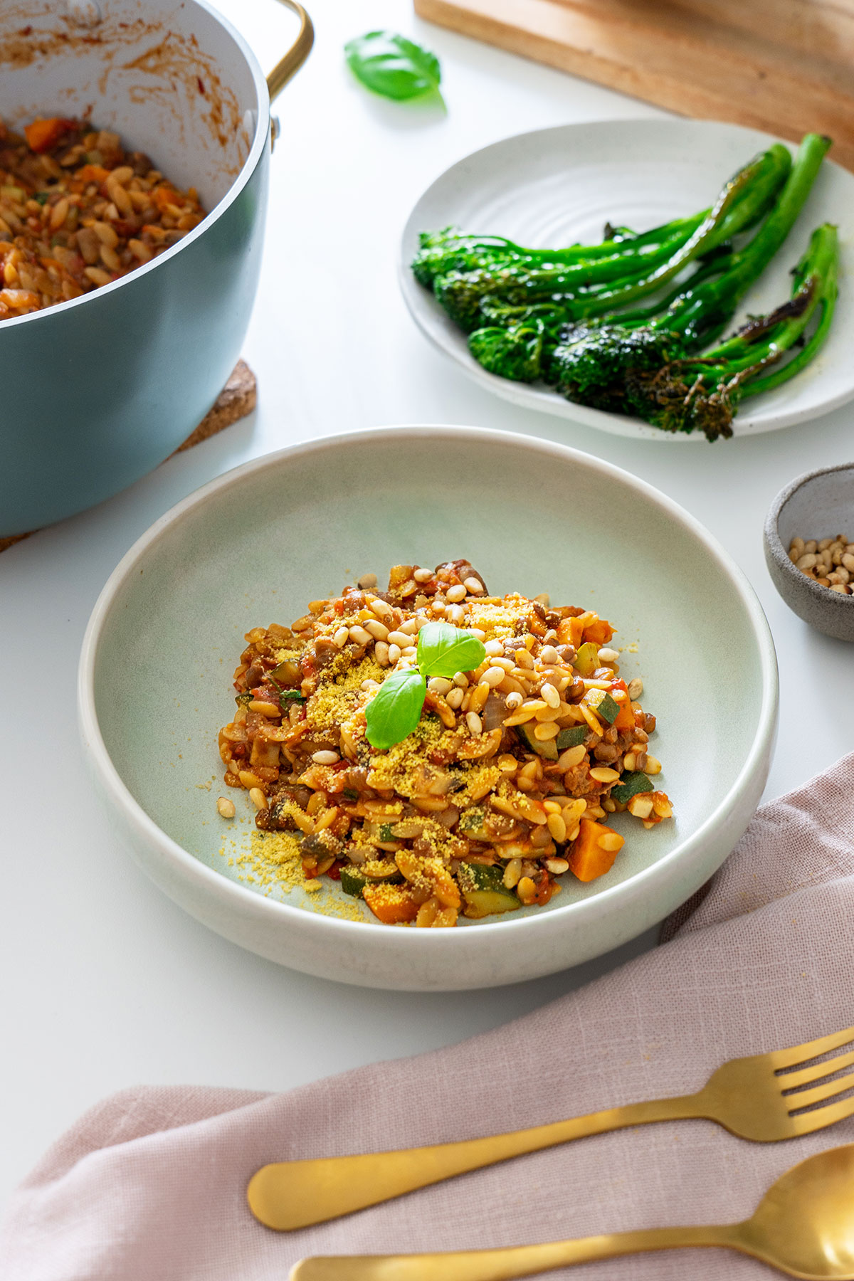 A bowl of vegan one-pot orzo Bolognese garnished with basil and pine nuts, served next to grilled broccolini.